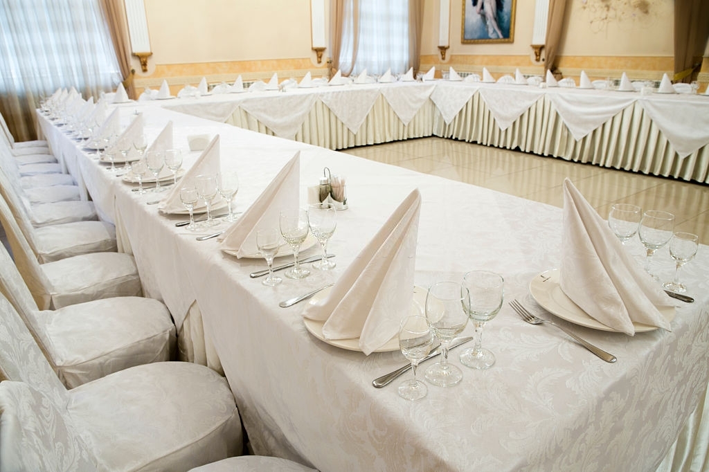 Beautifully organized event, glasses at served festive white tables ready.  Event in restaurant. Banquet, wedding decor, celebration. Catering and event. Wedding tables. Large restaurant hall.